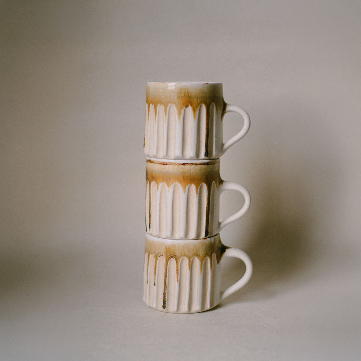 Three stacked ceramic mugs with a cream and brown drip glaze design, set against a neutral warm gray background.