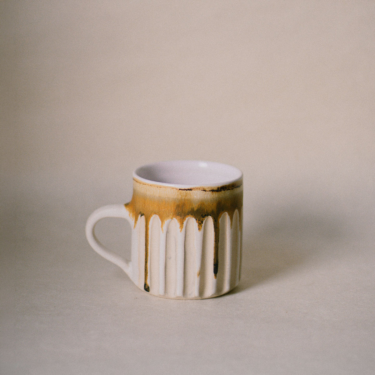 A ceramic mug with a ribbed fluted body, a white handle, and a dripping gold-brown glaze on an off-white base, set against a neutral beige background.
