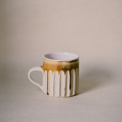 A ceramic mug with a ribbed fluted body, a white handle, and a dripping gold-brown glaze on an off-white base, set against a neutral beige background.