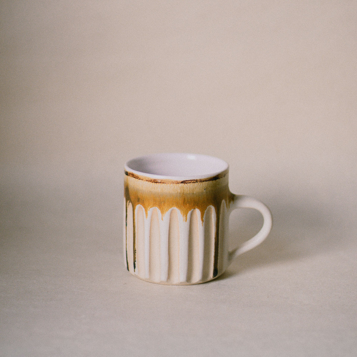 A small ceramic mug with a white ribbed body, a iron oxide drip glaze at the rim, and a white handle, set against a plain beige background.