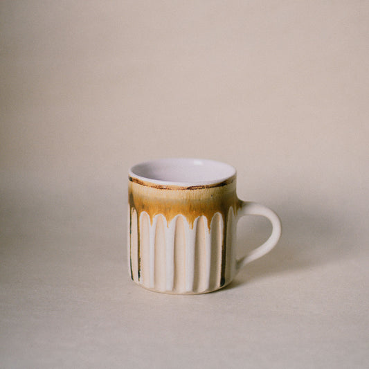 A small ceramic mug with a white ribbed body, a iron oxide drip glaze at the rim, and a white handle, set against a plain beige background.