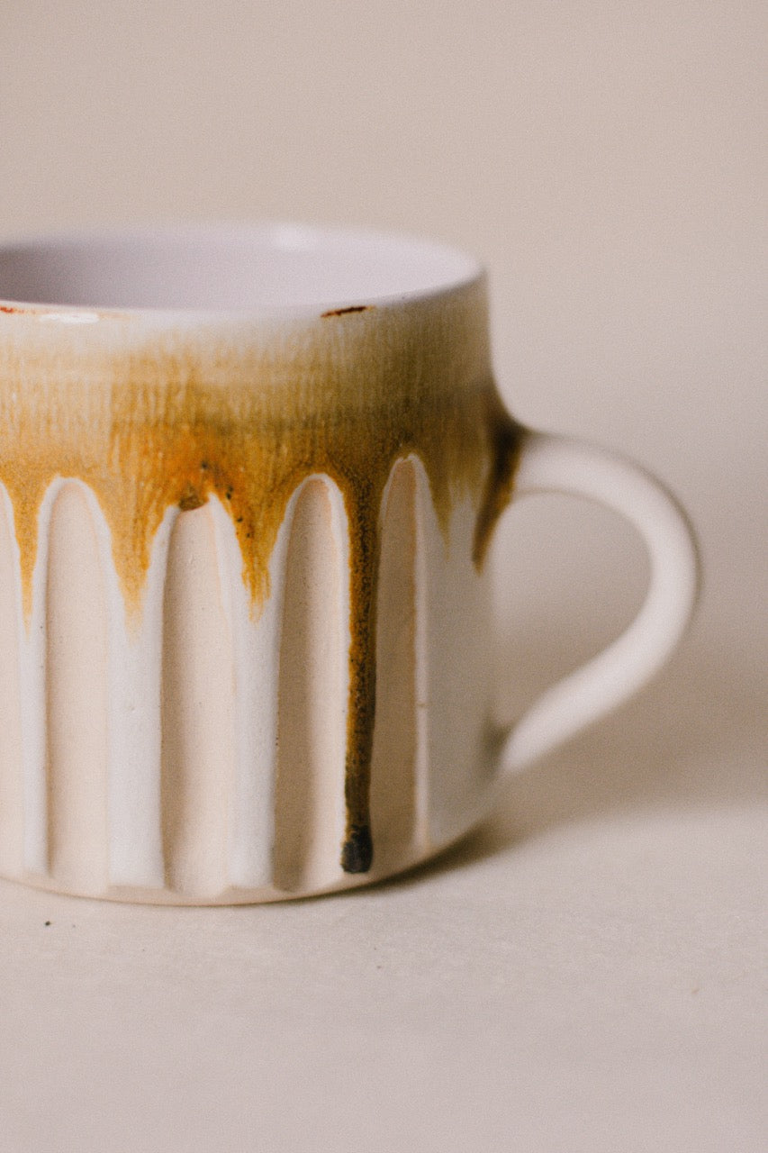 A close-up of a ceramic mug with a white ribbed body, a curved handle, and a glossy finish with a brown drip glaze design.