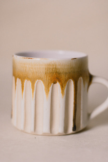 A ceramic mug with a white ribbed body, featuring a brown drip glaze design around the rim and handle, set against a plain beige background.