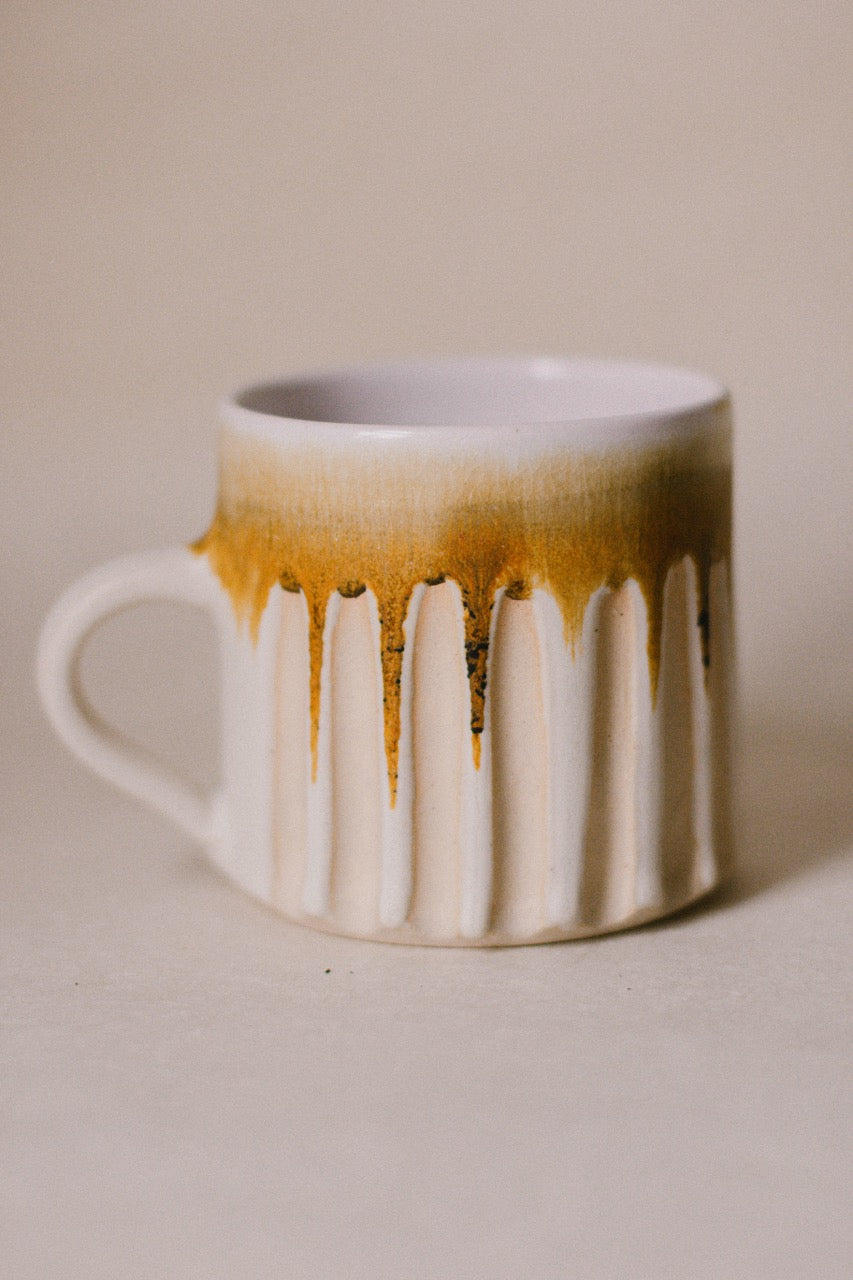 A white ceramic mug with a ribbed fluted design, featuring a glossy amber-brown drip glaze pattern around the upper half and handle, set against a neutral beige background.