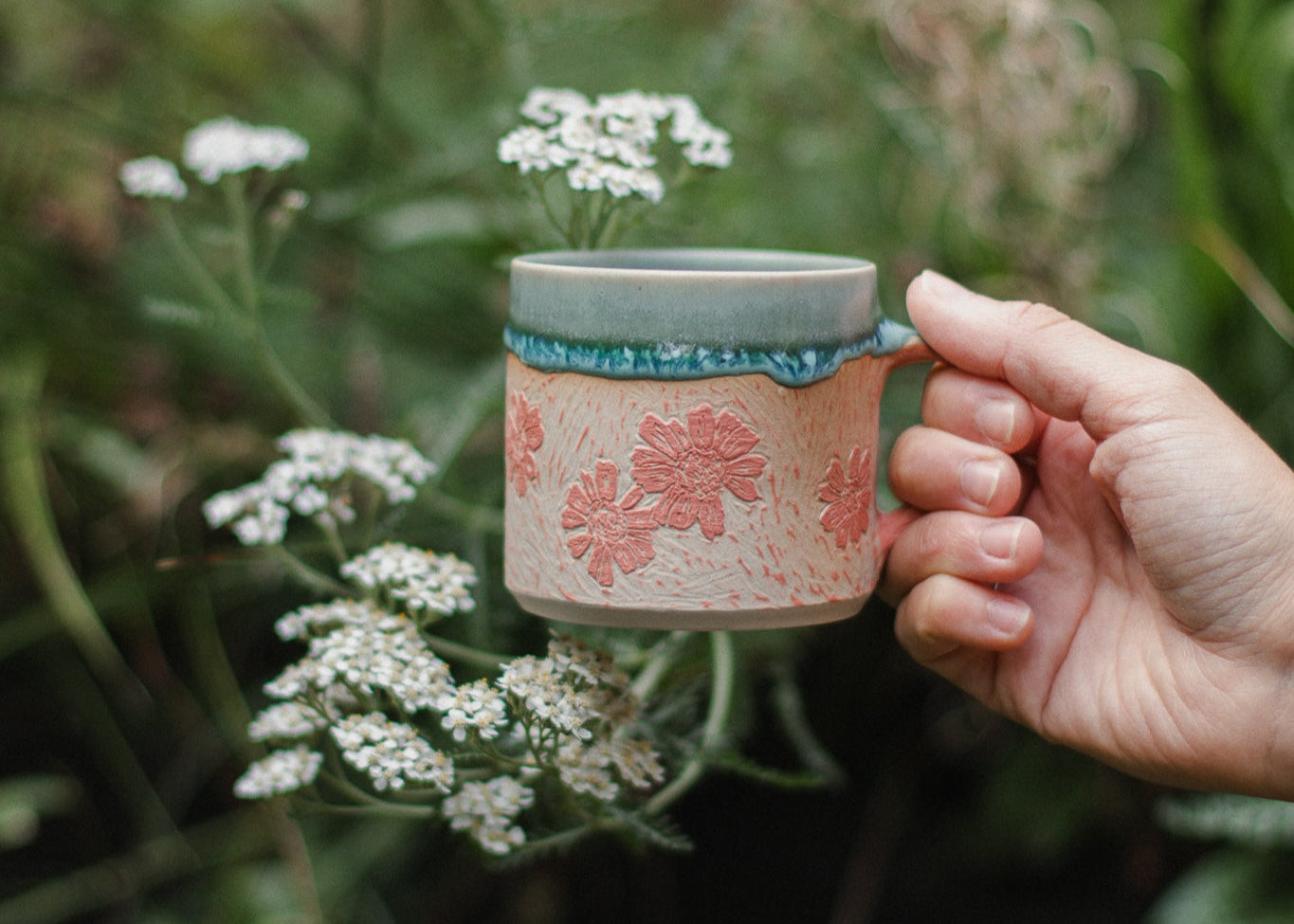 Hand holding a ceramic mug with floral design in a natural setting with flowers.